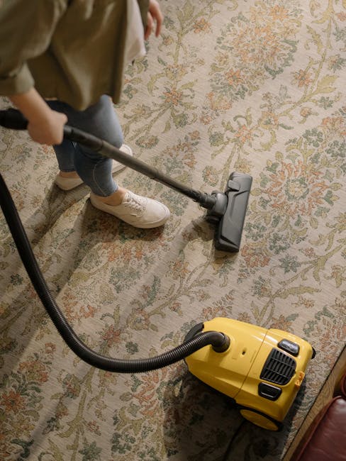 A man with dark, curly hair and a beard, wearing an orange sweater and grey trousers, is installing or patching a wall switch or outlet in a bright, white living room. The room has hardwood flooring, 