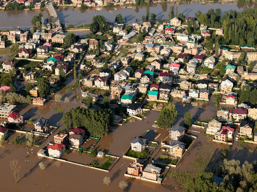 A flooded residential front yard during a heavy rainstorm, with muddy water covering the grass and pathway leading to a two-story house with a tiled roof and dormer windows. The house has a white exte