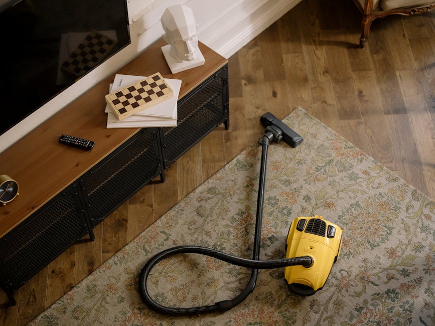 Close-up image of a vacuum cleaner head actively cleaning a patterned, textured carpet in a residential room. The carpet features a floral design with muted green, beige, and orange tones, and shows s