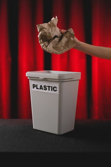 A person's arm is extended over a white plastic recycling bin labeled 'PLASTIC' with a black printed sign, positioned on a dark surface against a red curtain background. The person is holding a crumpl