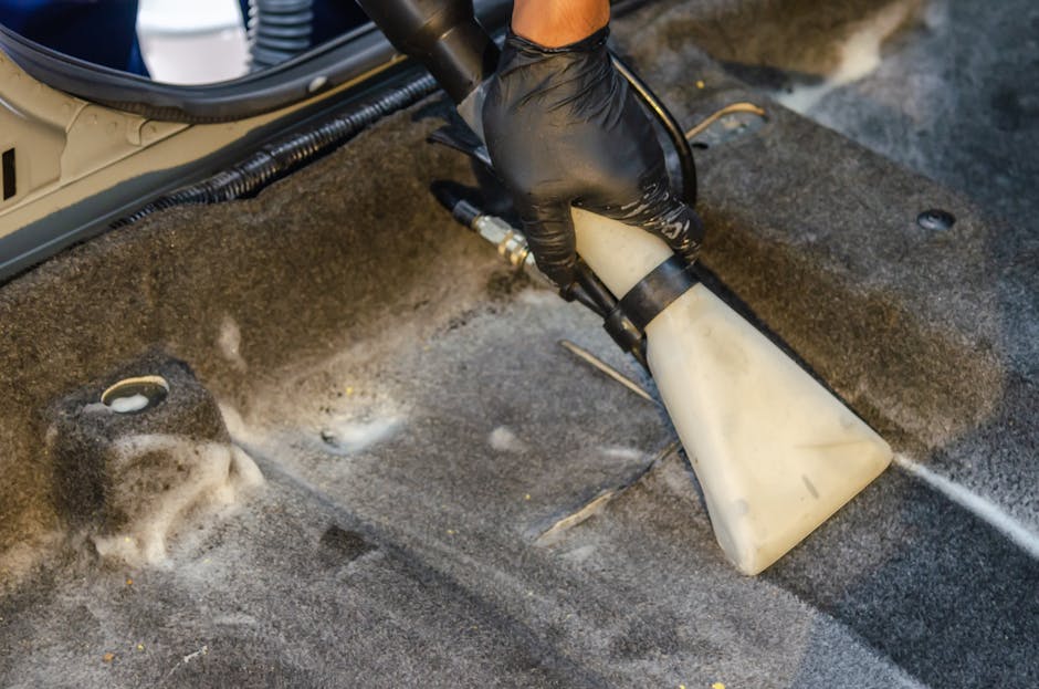 A person using a yellow vacuum cleaner with a black hose attachment to deep clean a patterned, beige and green floral area rug in a domestic setting. The individual is wearing beige shoes, grey jeans,