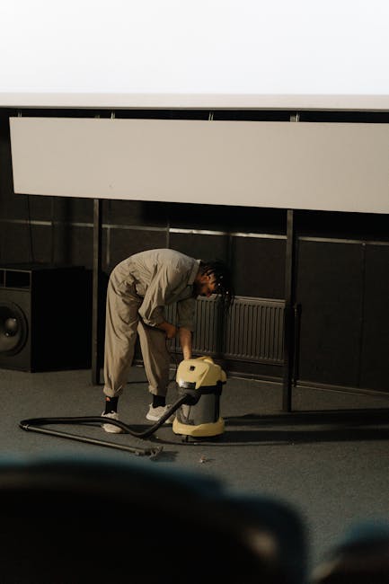 A close-up view of a person using a vacuum cleaner with a black and grey head and a long, black handle to clean a patterned area rug on a well-lit wooden floor. The rug features intricate floral desig