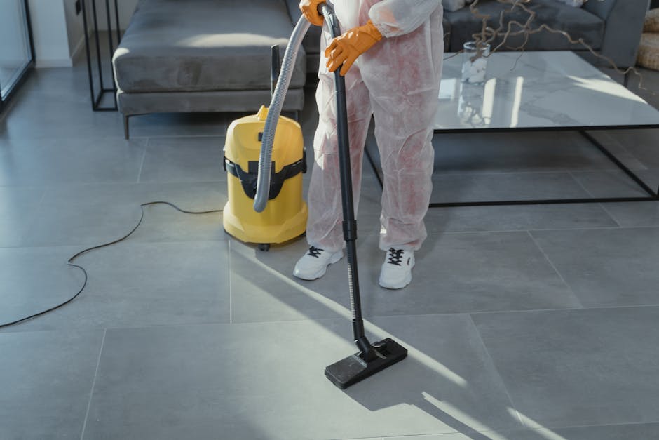 A professional cleaner wearing white coveralls and orange gloves is performing surface cleaning on a large gray tiled floor in a modern living room using a vacuum cleaner with a long black hose and a